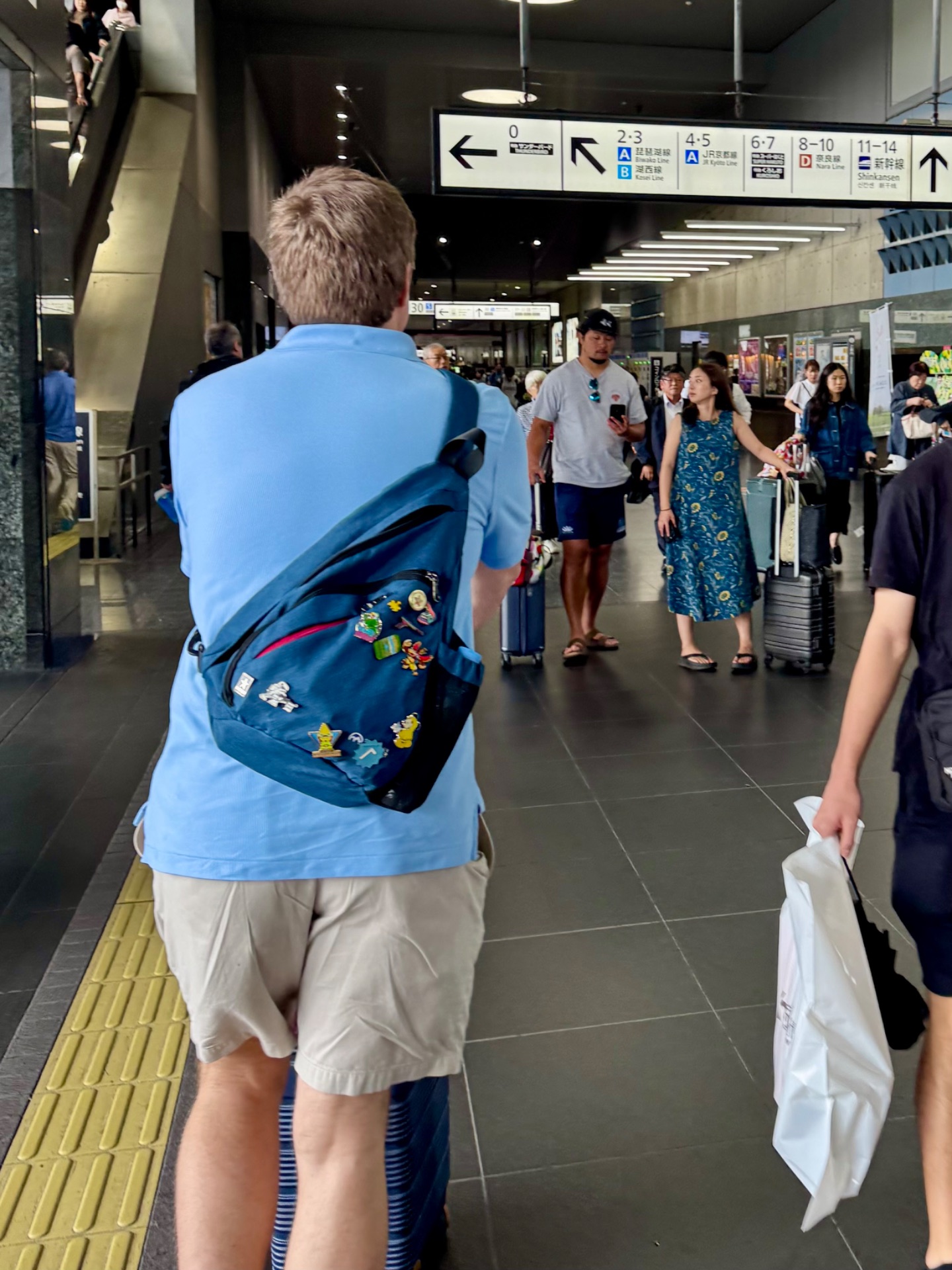 Man with Pokemon backpack walking through modern subway station with other travelers and directional signage overhead.