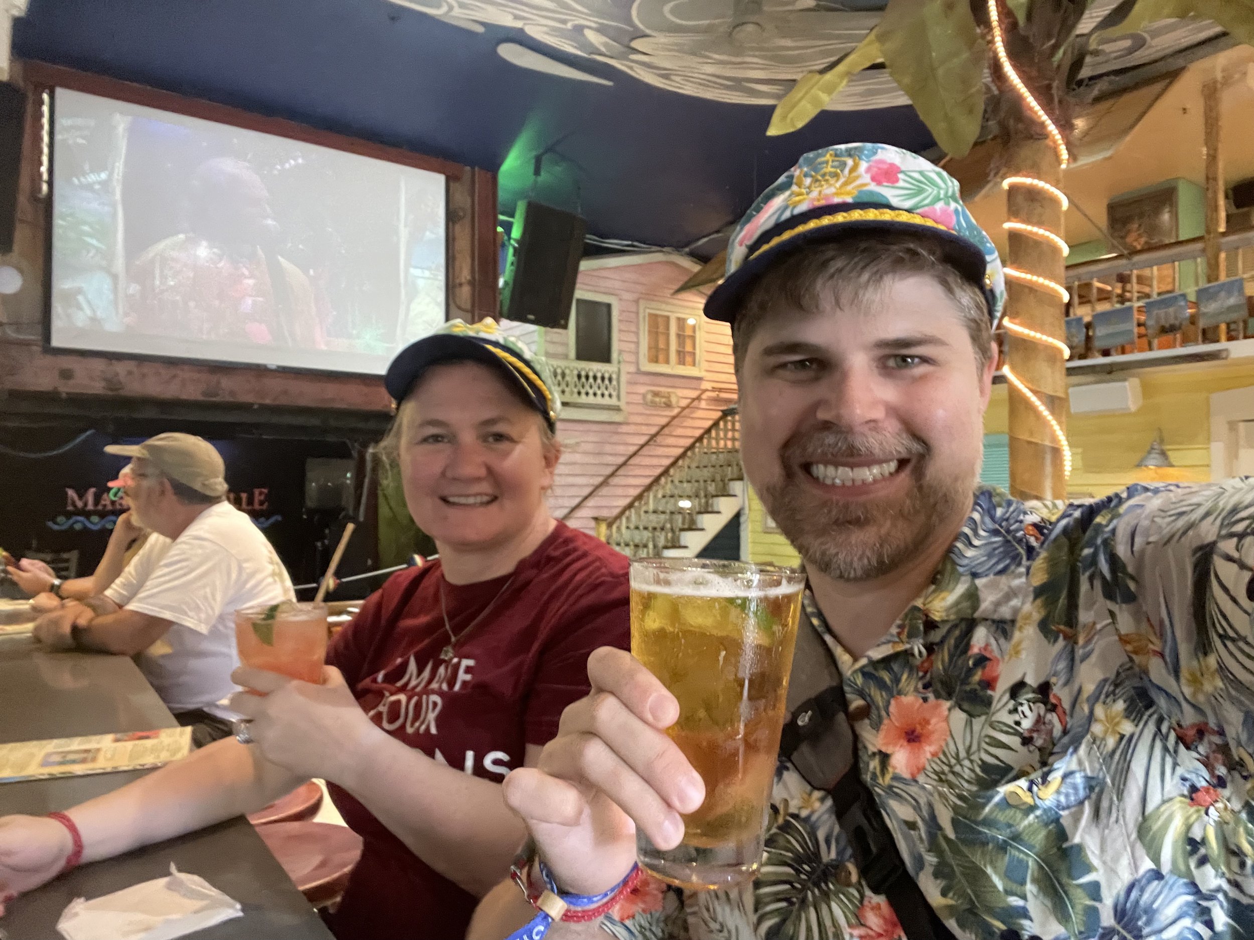 Two people wearing colorful caps holding drinks at cruise ship bar