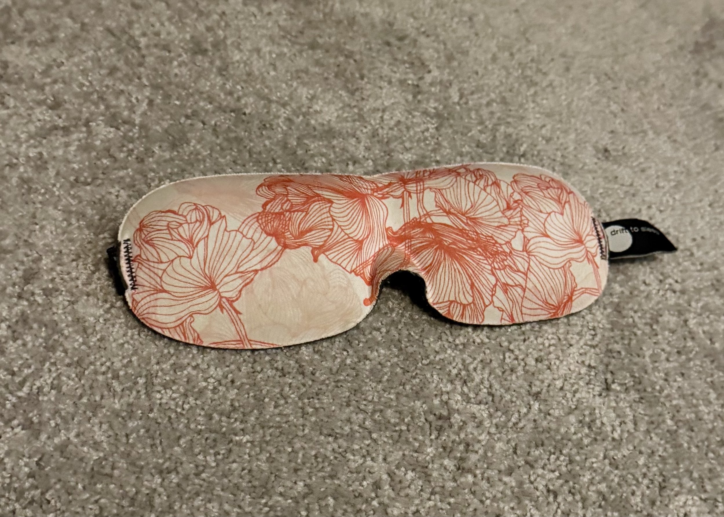 Sleep mask with coral and red floral line drawing pattern on cream background, displayed on gray surface.