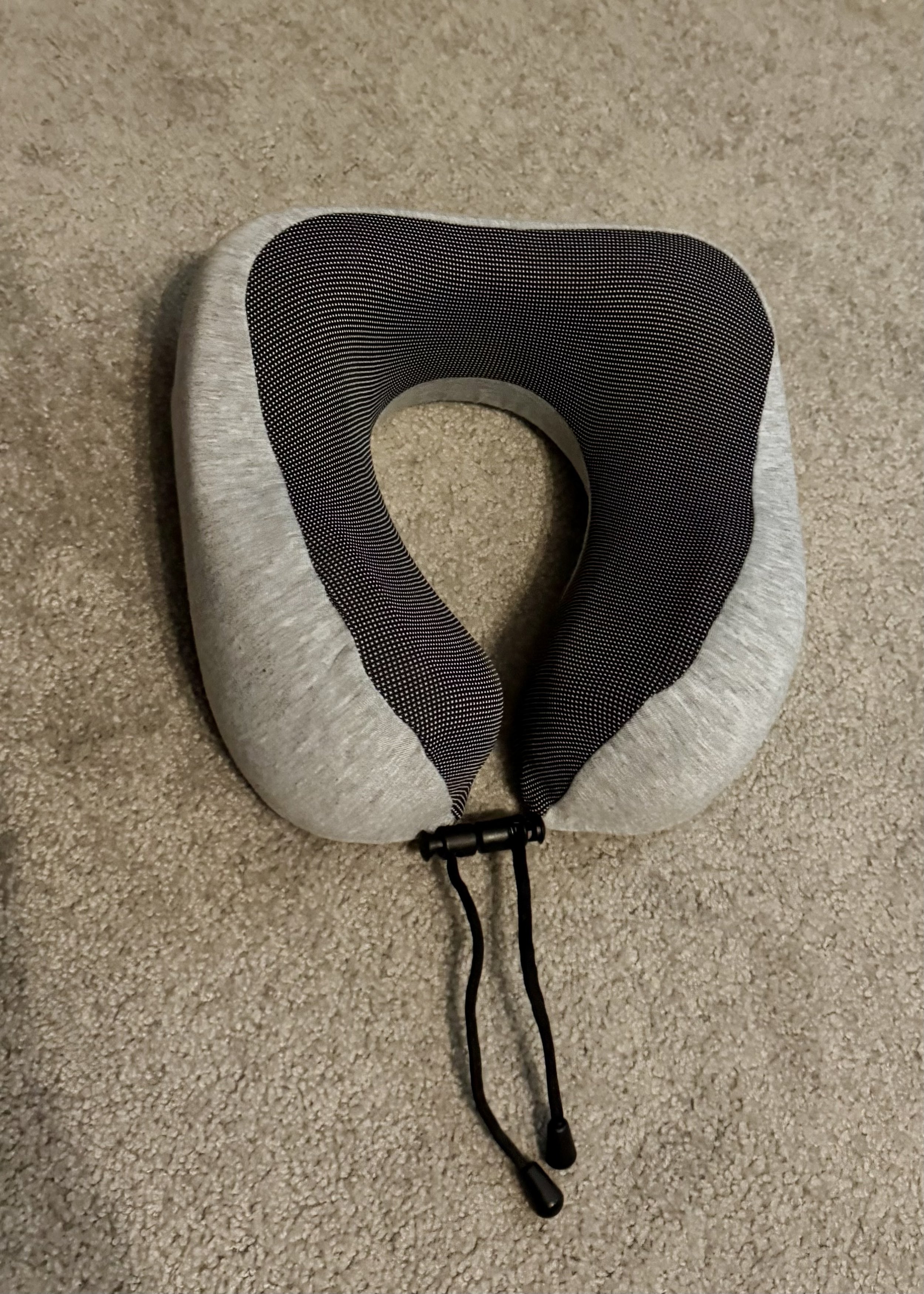 Gray memory foam travel neck pillow with dark mesh center and black drawstring cord on beige carpet.