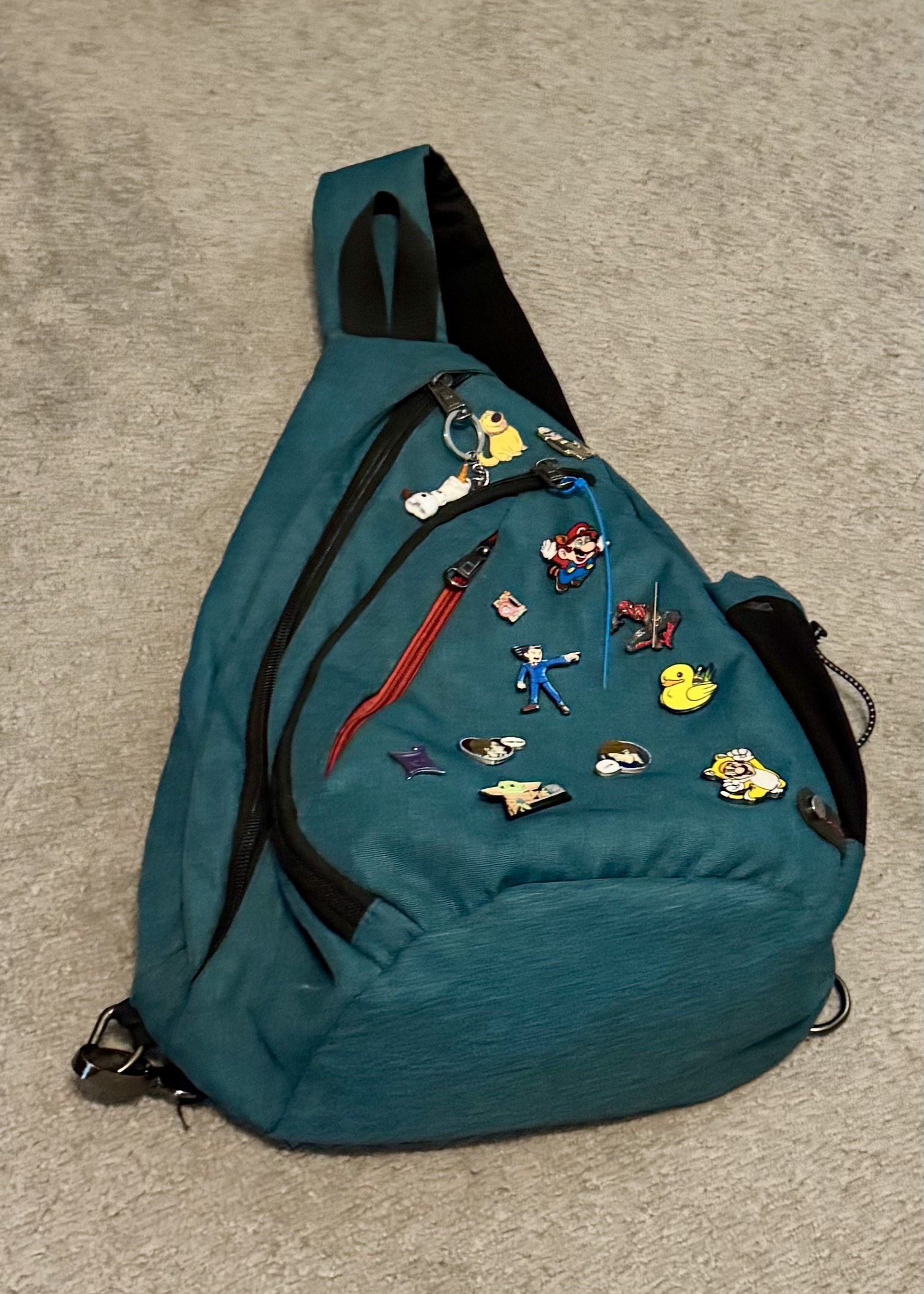 Teal sling backpack decorated with enamel pins featuring pop culture characters and icons.