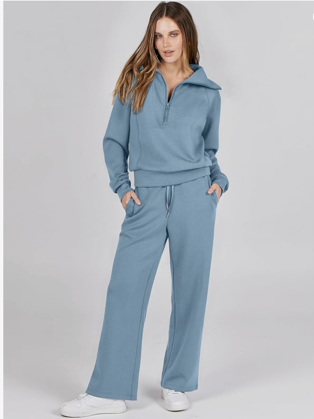 Woman wearing blue tracksuit with half-zip hoodie and matching sweatpants, styled with white sneakers against neutral background.